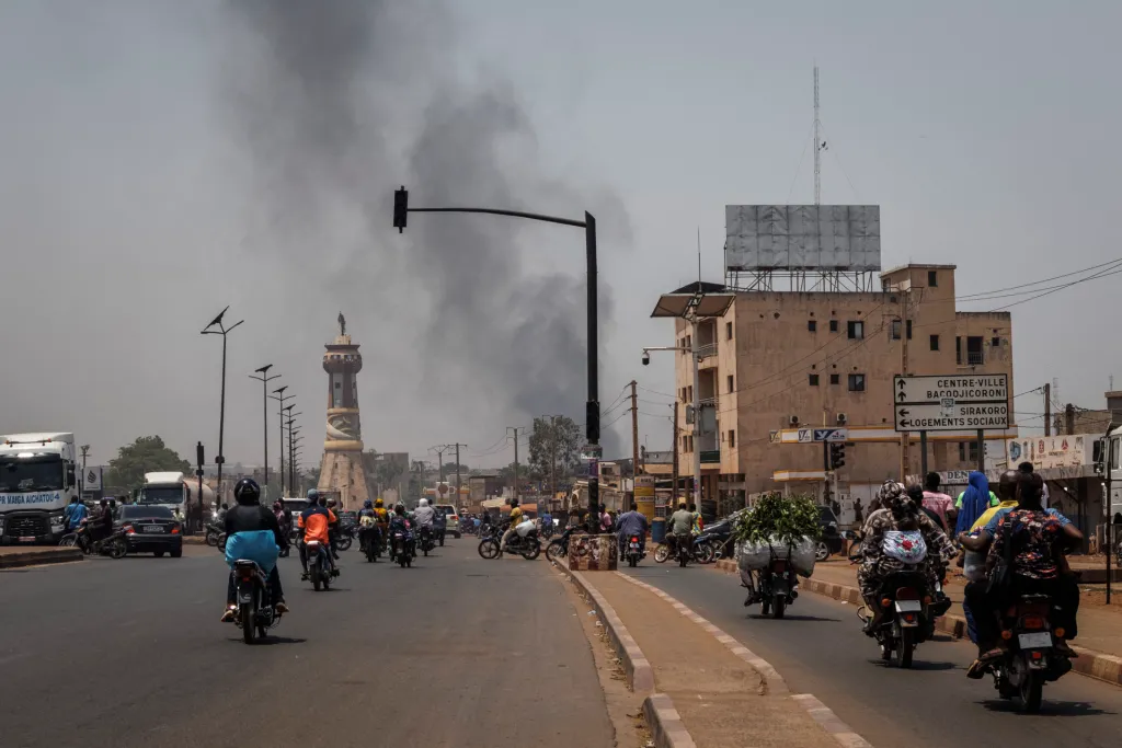 A column of black smoke rises above buildings as traffic, mostly motorcycles, pass in the foreground.